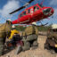 Four people in hard hats, yellow shirts, and green pants, kneel in the dirt as a red helicopter lands nearby
