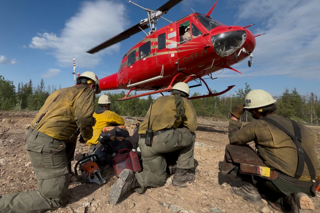 Four people in hard hats, yellow shirts, and green pants, kneel in the dirt as a red helicopter lands nearby